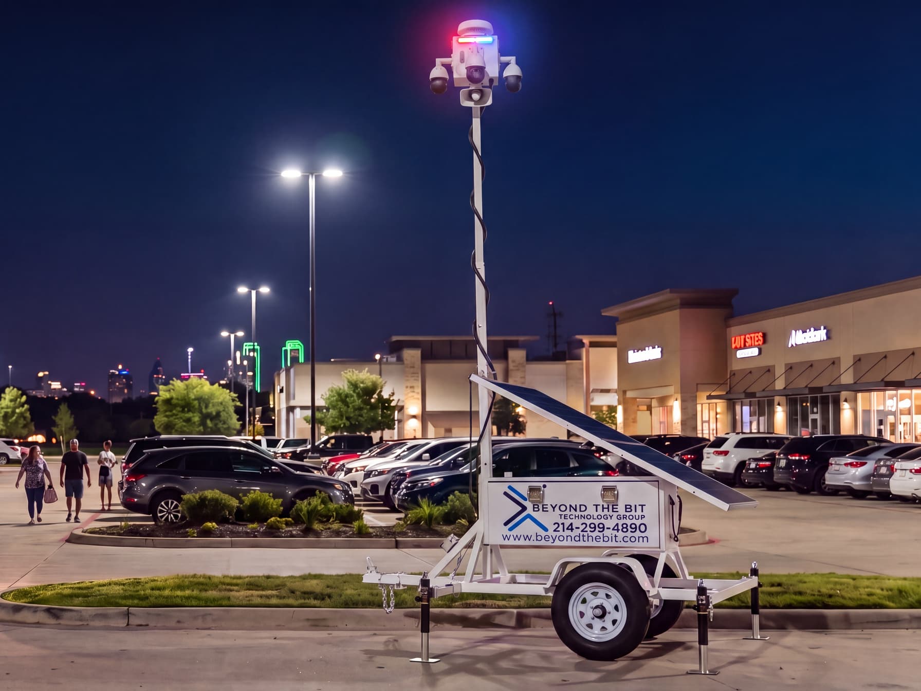 A Beyond the Bit security trailer with its mast raised at the edge of a busy Dallas-area retail plaza at night, storefronts lit and shoppers walking to their cars.