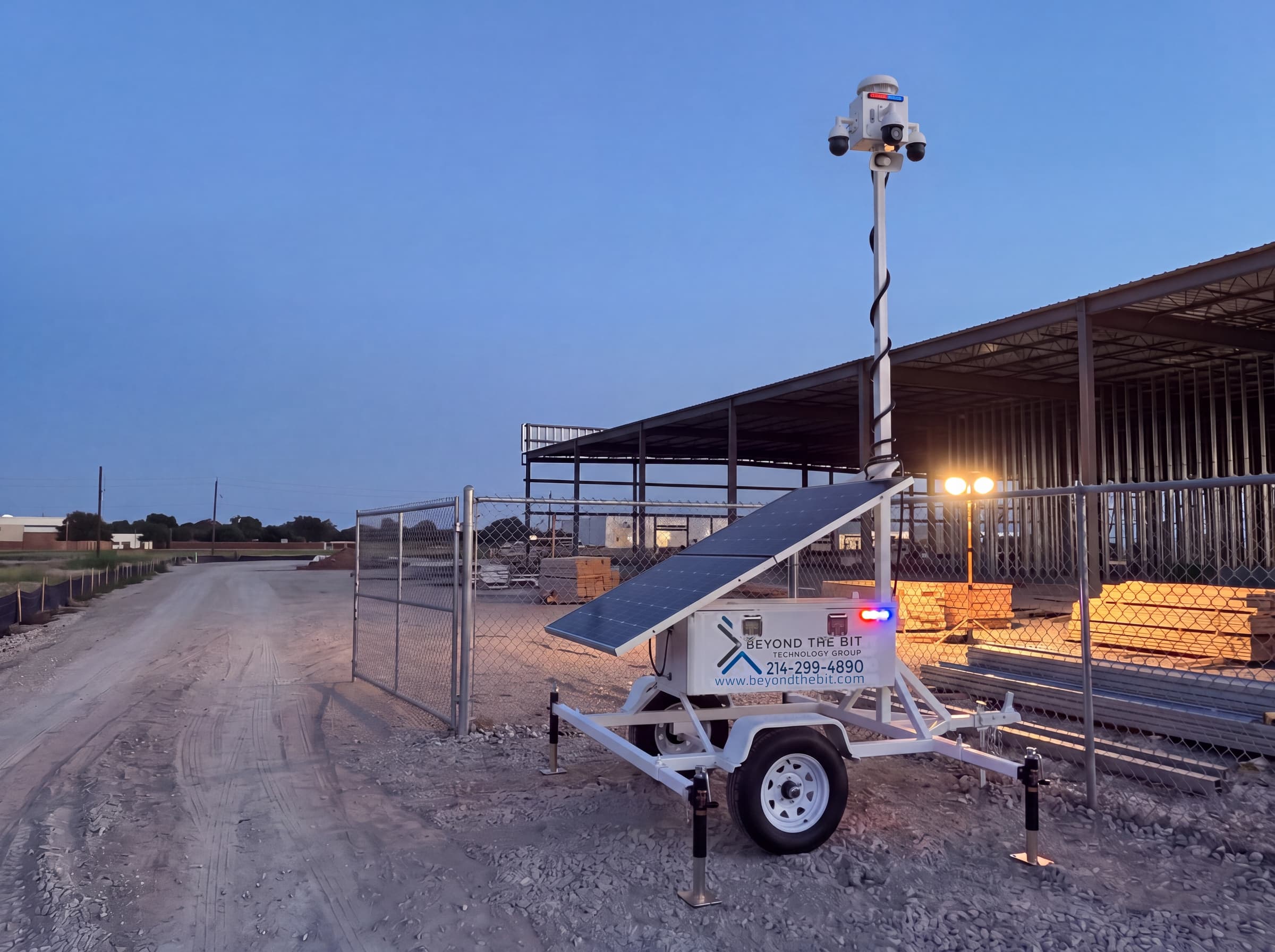 A Beyond the Bit security trailer parked on the shoulder just inside a fenced construction site entrance at dusk, camera mast raised, the open vehicle gate and drive lane clear beside it.